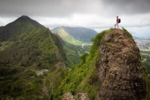 A man stands triumphantly on a mountain peak, gazing over a vast, scenic valley below him.