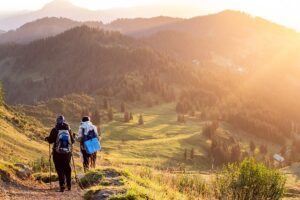 Three hikers with backpacks ascend a mountain trail, surrounded by lush greenery and rocky terrain.