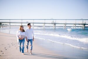 A pregnant couple strolls hand in hand along the beach, with gentle ocean waves lapping at the shore.